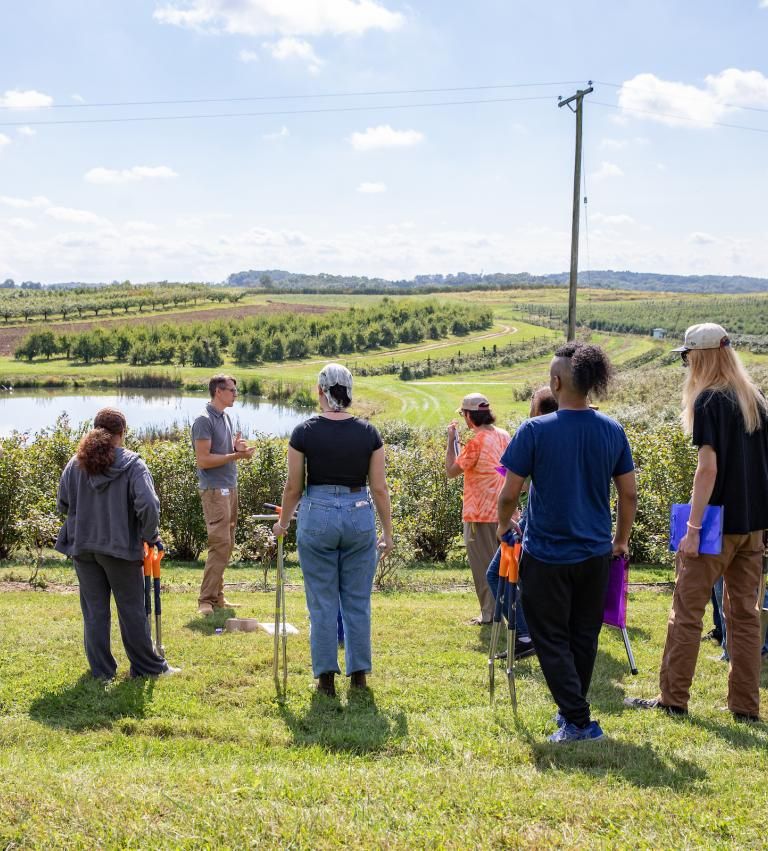 A group of seven students look on as their professor speaks to them in front of the open landscape of Shaw Orchard. Rows of plants are visible in the background.