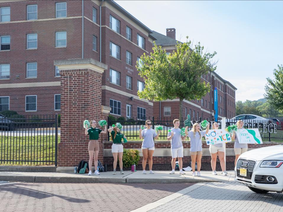 A group of student leaders hold signs and pom poms as they cheer for cars driving into the campus entry.