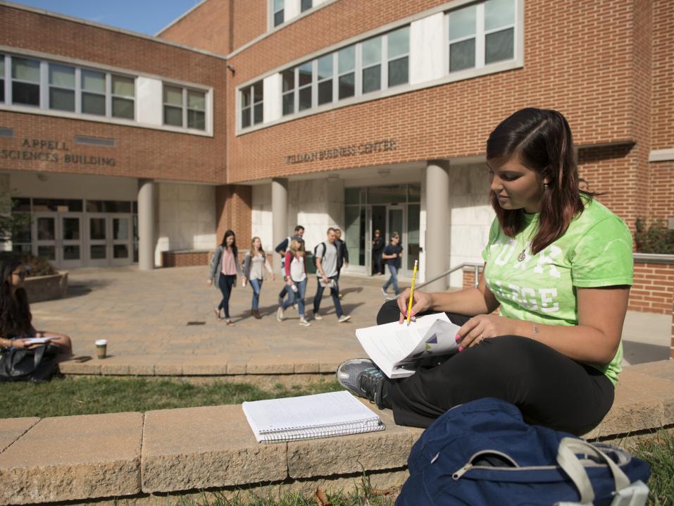 A student is outside in front of Willman Business Center and Life sciences writing in a notebook