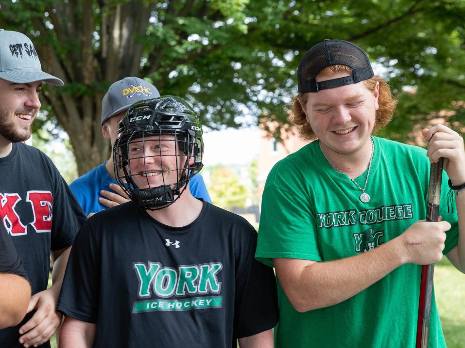 Four members of the ice hockey club laugh at an involvement fair outdoors. One wears a hockey helmet, while others hold hockey sticks.