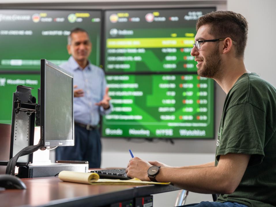 A professor and finance student in a finance classroom with a computer and large screen.