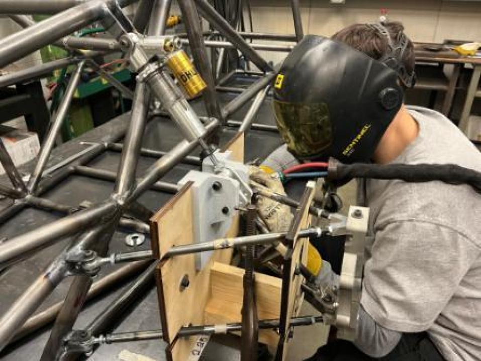 A student wearing welding equipment works on the frame of a car.