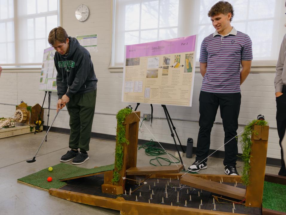 Students demonstrate a student-designed miniature golf course during a college engineering project showcase, with a poster presentation and hands-on testing of the course.