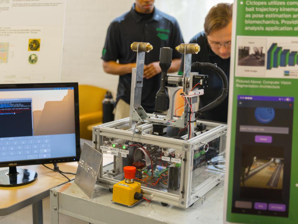 Students demonstrate an engineering project featuring a mechanical device, computer monitor and research poster.