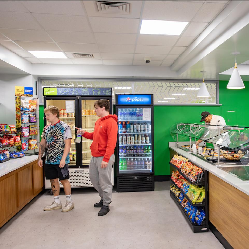 Two students shop in the convenience store section of Spart's Den while Chef Hector prepares food behind the serving station.