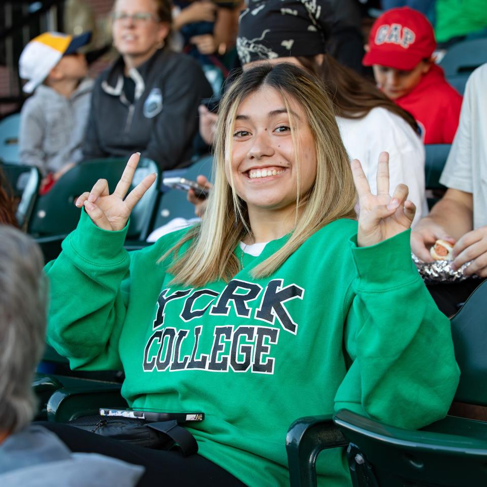 A student gives a peace sign to the camera and smiles from her seat at Revs Stadium during York College night.