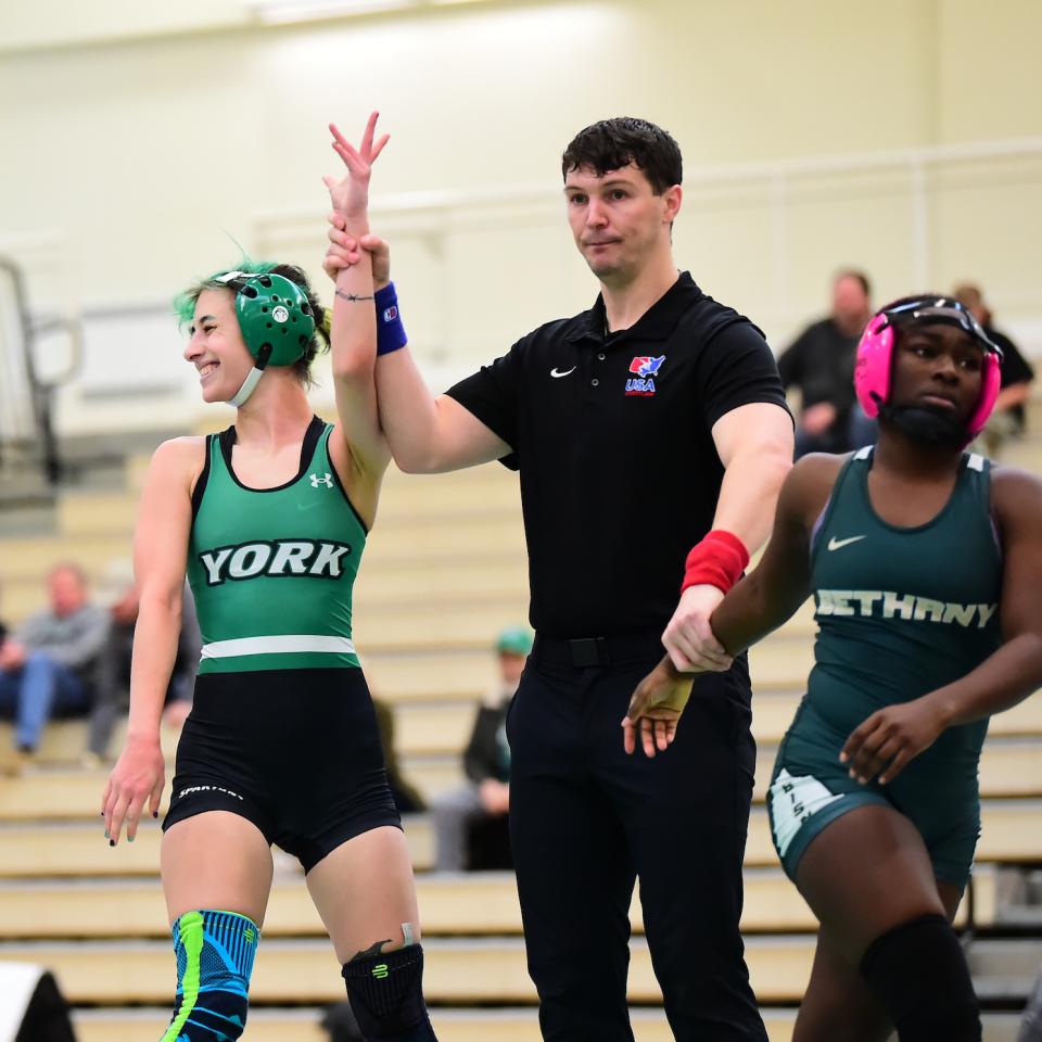 A YCP Women's Wrestler stands with a ref and her opponent, with her hand held in victory.