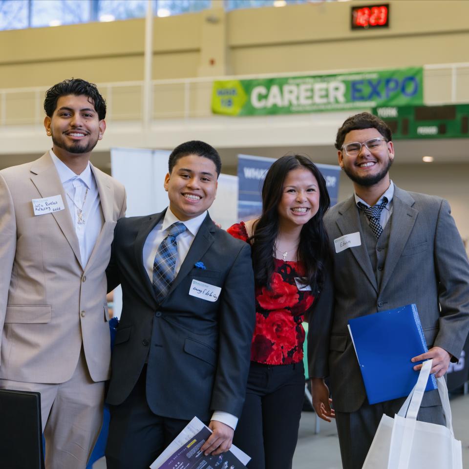 Four students stand together in dress clothes with a banner behind then that reads Career Expo.
