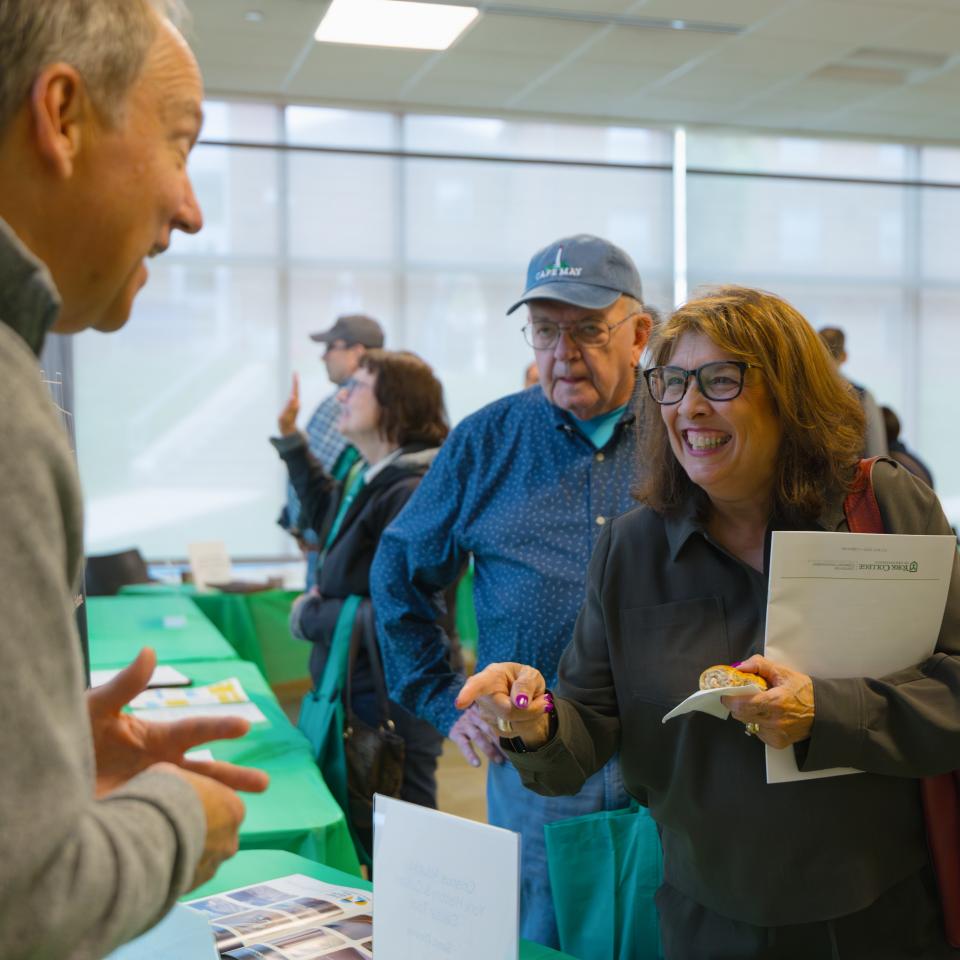A room full of people and a man and a women talk to someone behind a table with a green tablecloth.