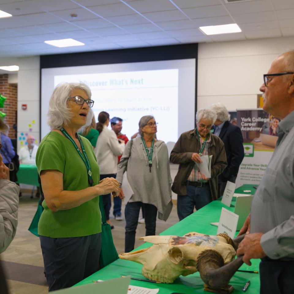 A room full of people and a women talks to someone behind a table with a green tablecloth.