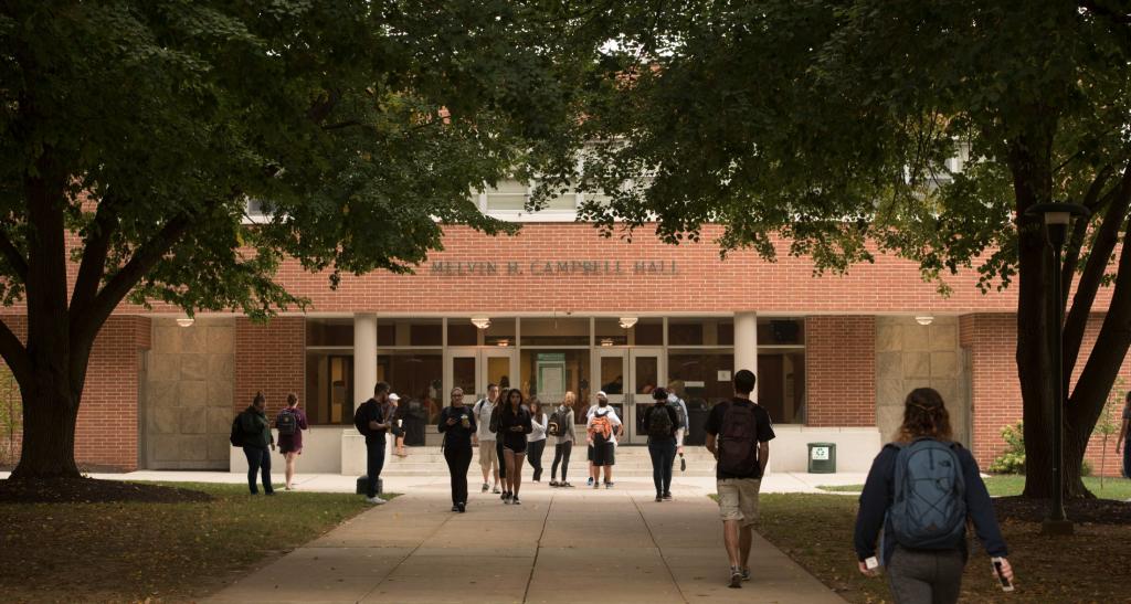 Student walking outdoors in front of Melvin Campbell Hall, a large red brick building surrounded by trees.