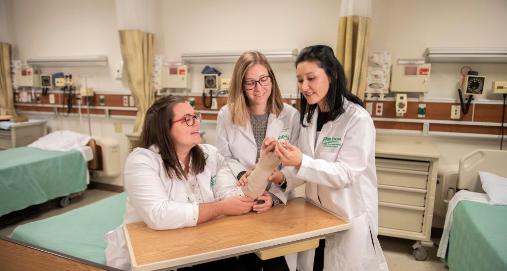 Two nurses in white lab coats talk to patient as an ace bandage is wrapped around her wrist in a clinical environment.