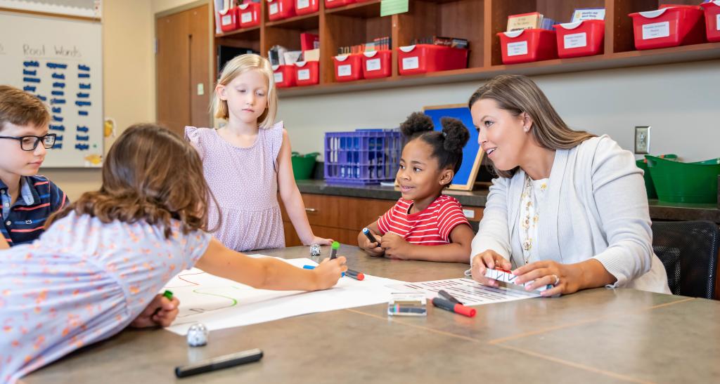 A teacher sits at an elementary school classroom table with four young students. Markers and paper are spread out in front of them.