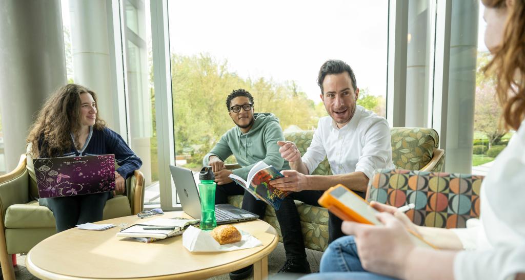 A professor and three students chat in a sitting area on campus.