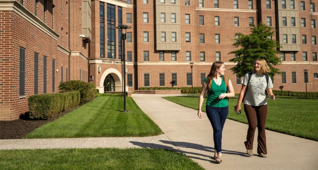 Two students carry backpacks as they talk and walk in front of the tall brick front of Northside Commons' buildings.