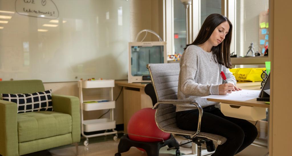 A student studies at a desk in a workspace with a whiteboard, a lounge chair, and windows.