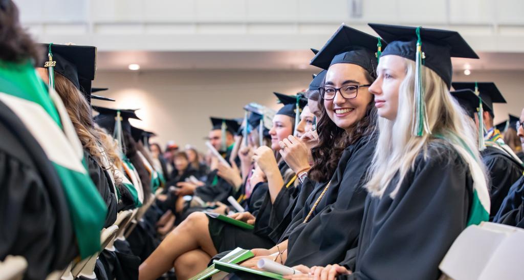 A student dressed in cap and gown beams at the camera while seated for her commencement ceremony.