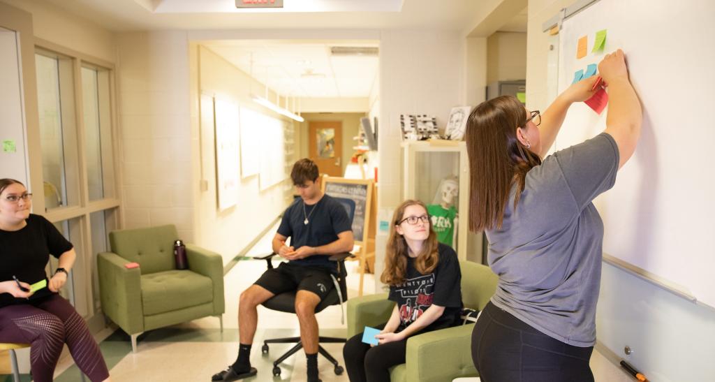 A student places post-its on the whiteboard while three other students, seated, look on.