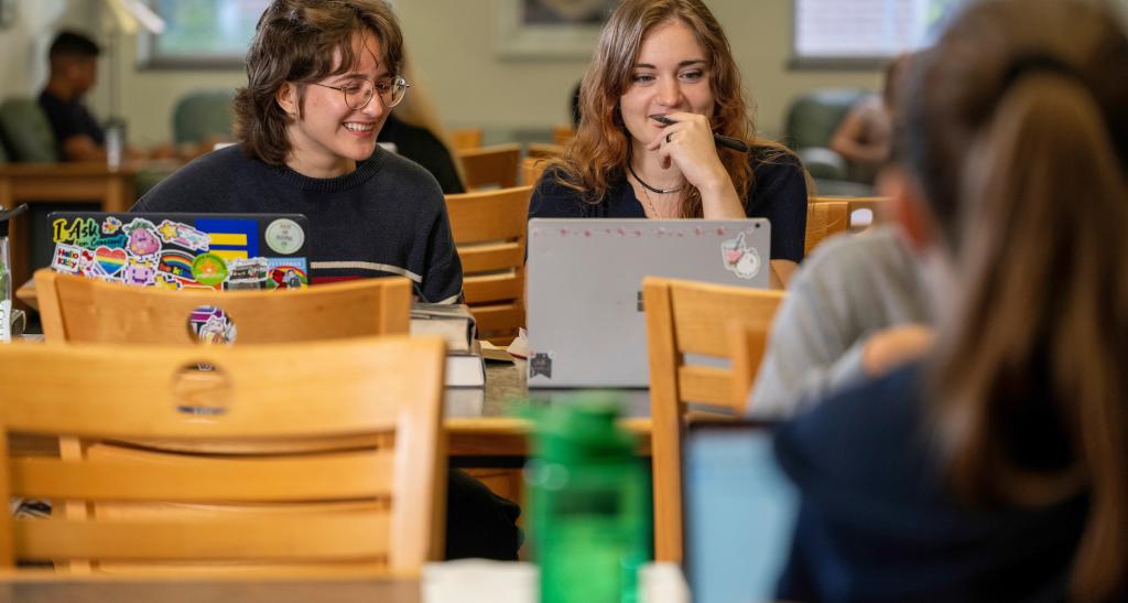 Two students studying at a table in the library with notebooks and a laptop