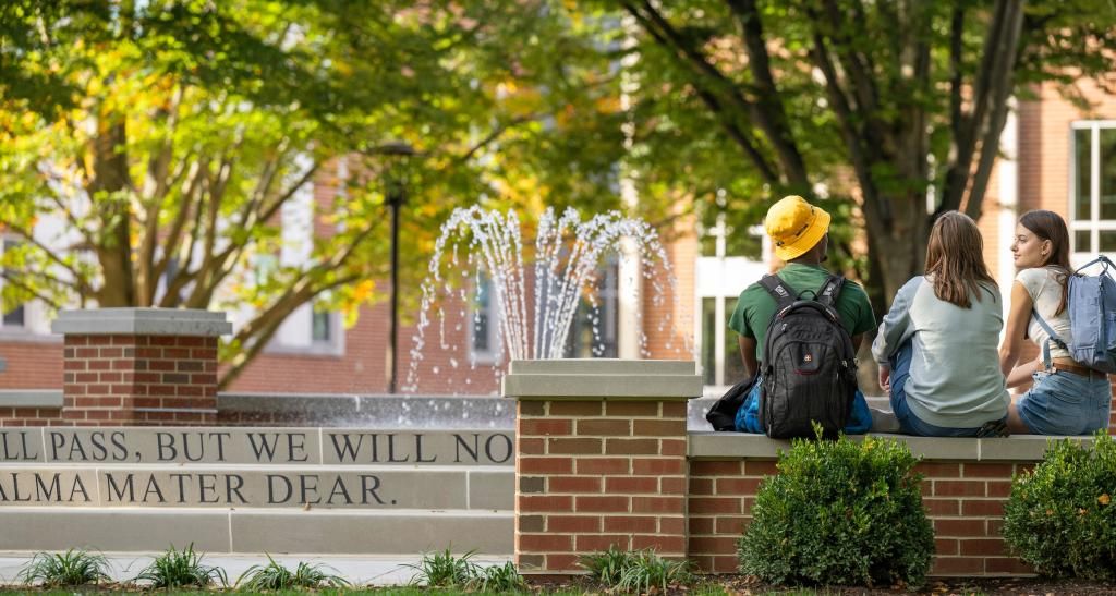 Three students sit on a half wall beside a fountain in the campus quad, their backs to camera.