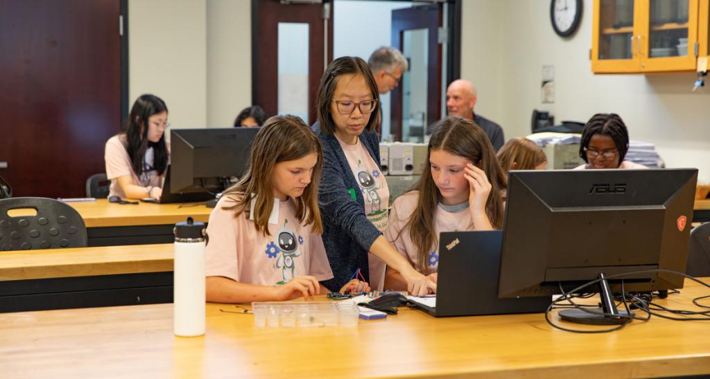 Two girls in the EmpowerHer steam program sit at a desk with a teacher behind them pointing something out