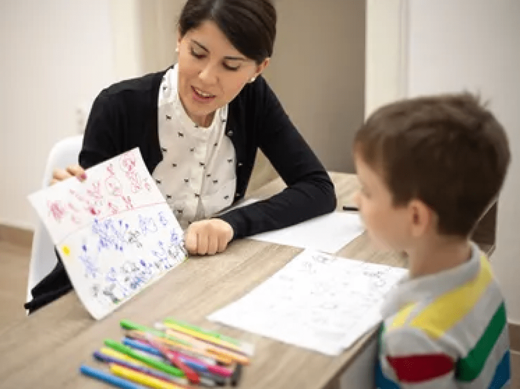 A teacher holds up a child's drawing as the child looks on from his seat across a desk covered in markers and paper.