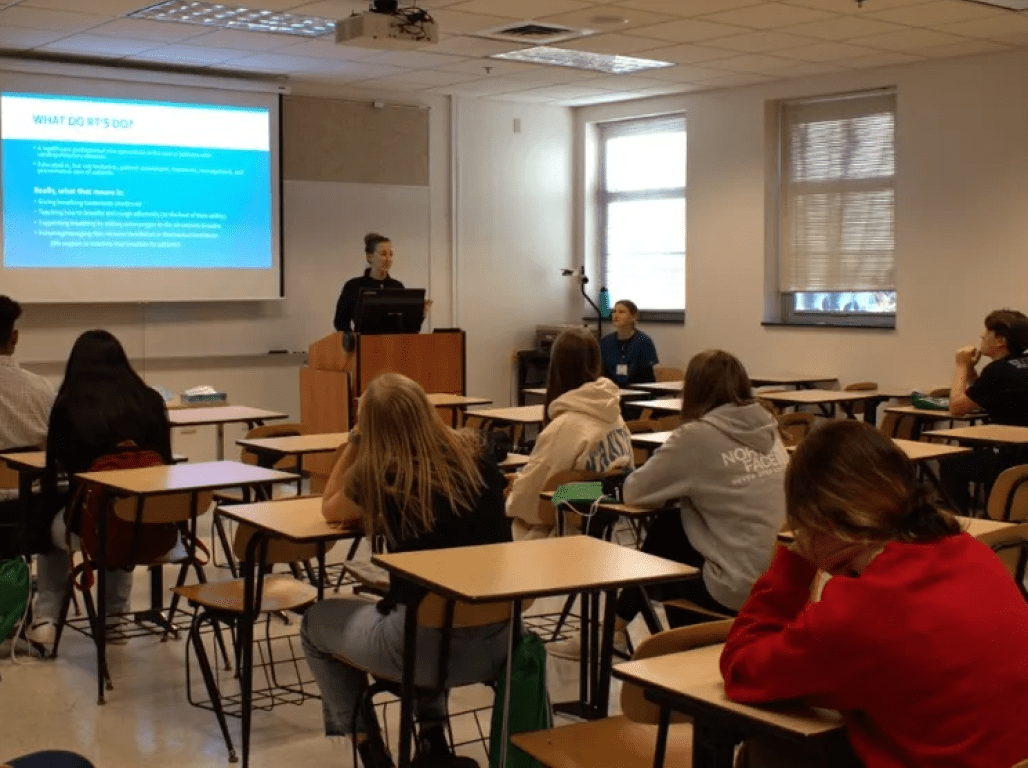 A group of high school students listen to a professor in a classroom.