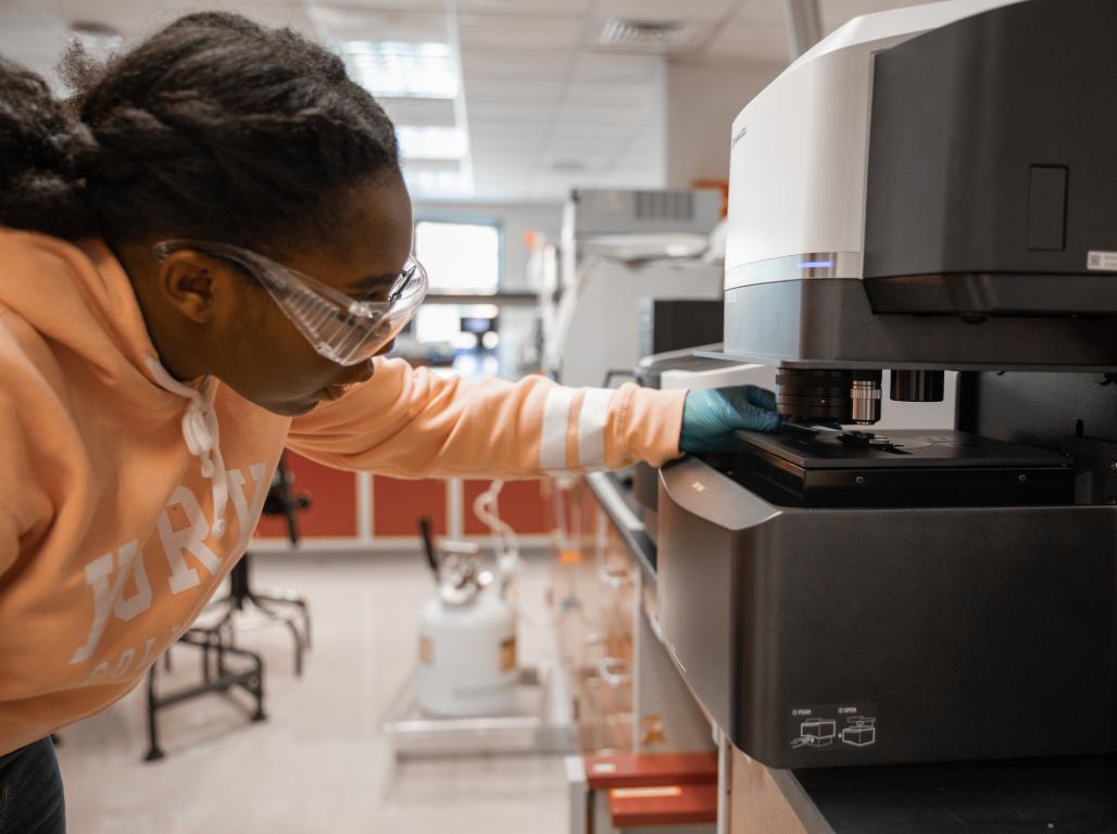 A student wearing lab safety goggles and gloves works with equipment in the chemistry lab.