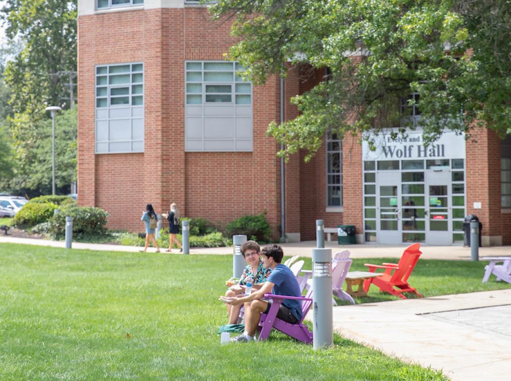 Two students sitting in front of Wolf Hall on Adirondack chairs.