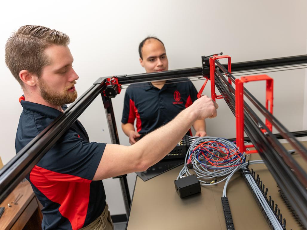 Evan Lehr and Marcos Rivera wear uniform polos and work on engineering equipment.