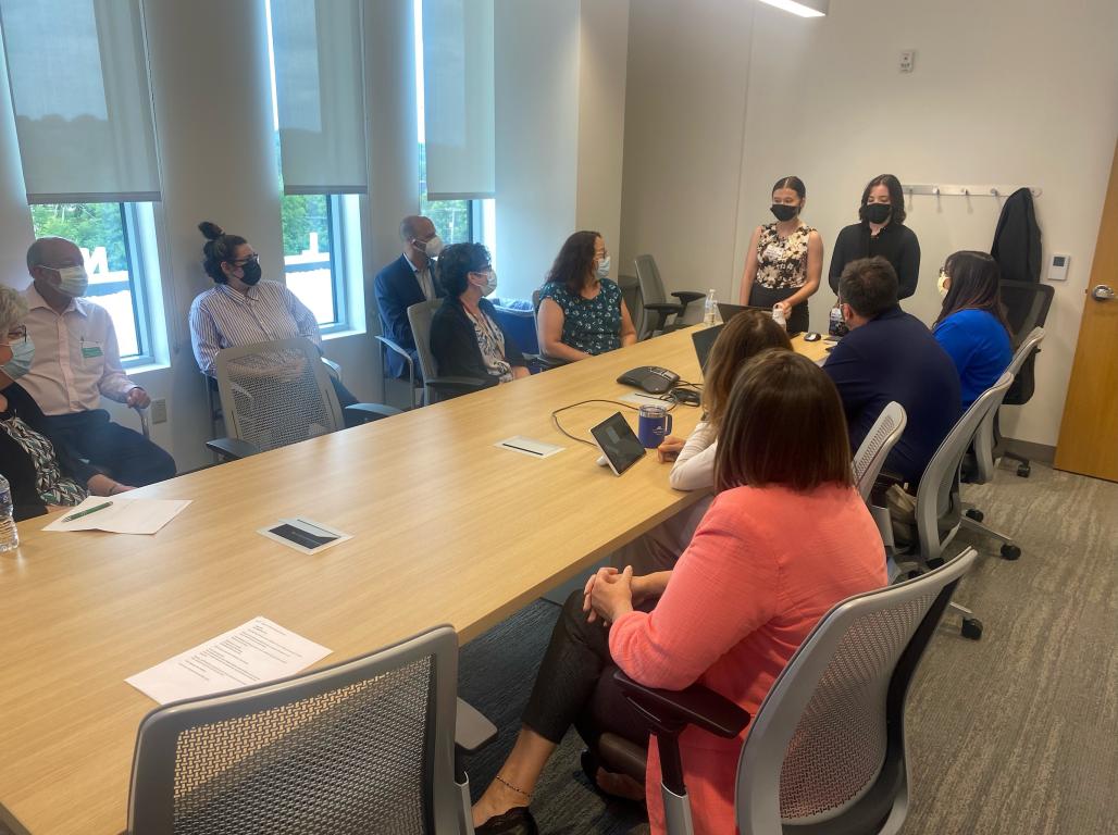 Two students present from the head of a conference room table while several professionals look on.