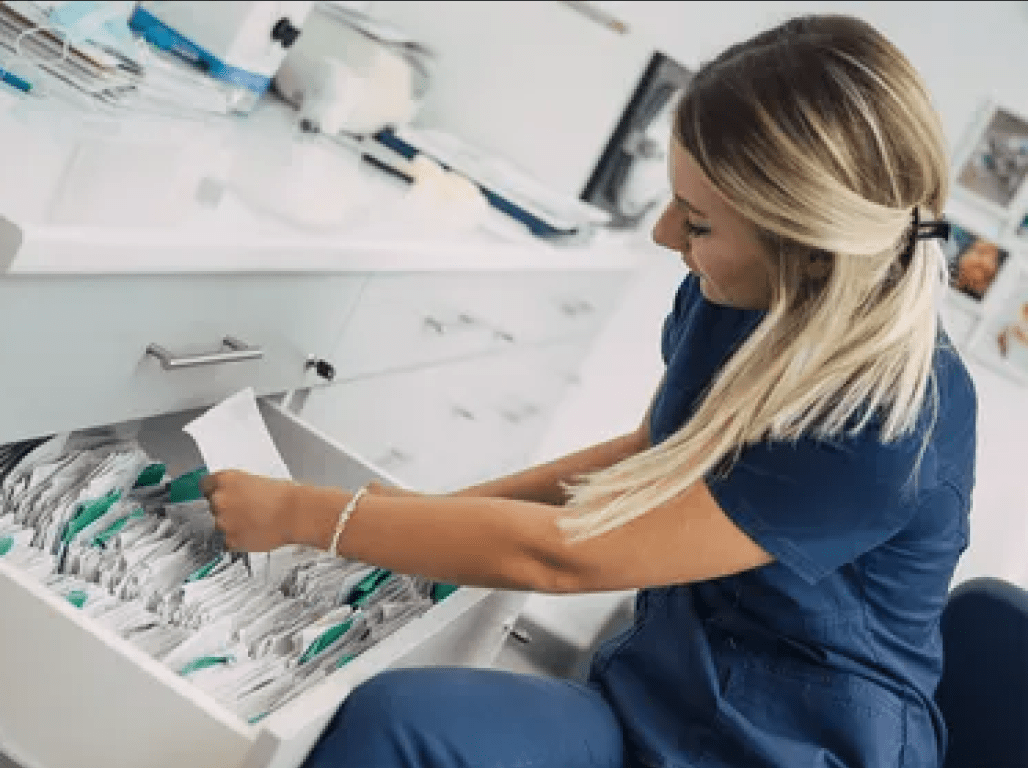 A health professional wearing scrubs looks through a filing cabinet in a medical office.