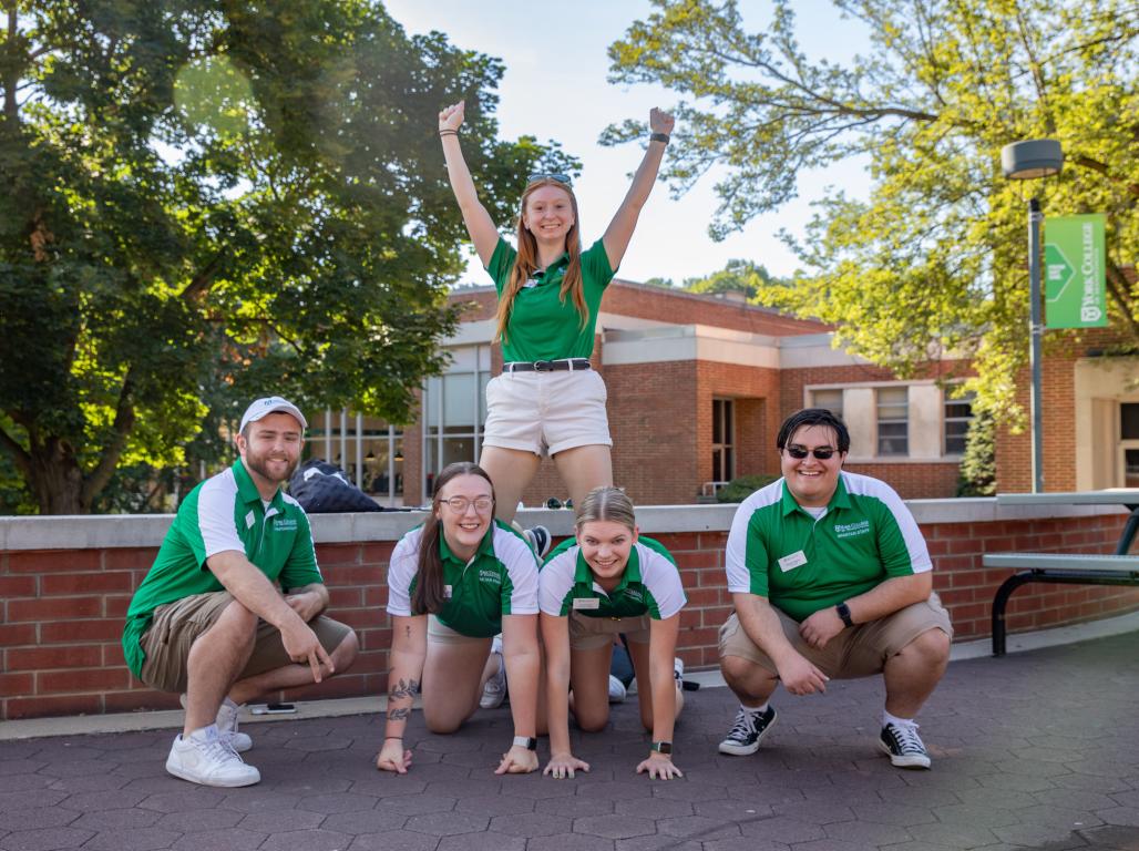 Four students pose for a group photo. They smile while making a human pyramid; the student on top of the pyramid throws her hands in the air.