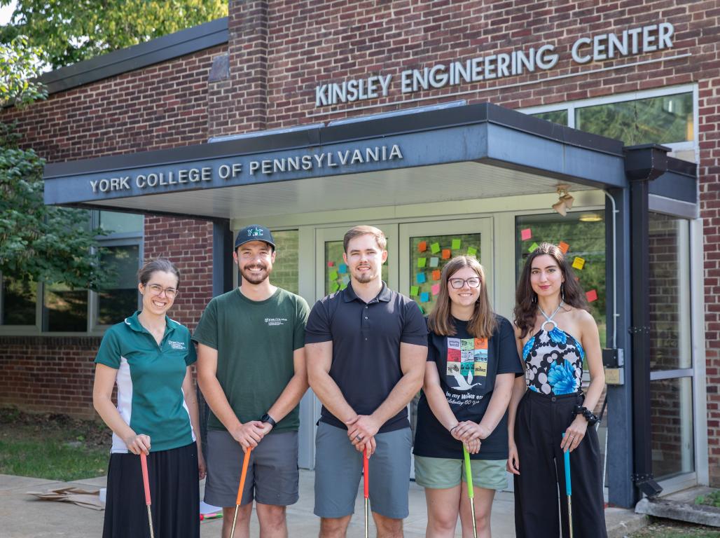 Dr. Ashley Earle and four students from her Intro to Engineering class pose for a group photo in front of the Kinsley Engineering Center; they hold mini golf clubs in their hands.