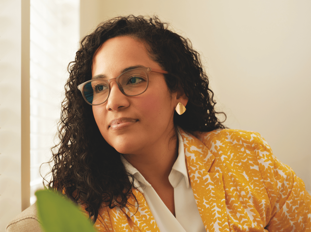Thaís Carrero looks out a window from a comfortable office environment. She sits in a lounge chair, and a houseplant is visible in the foreground.