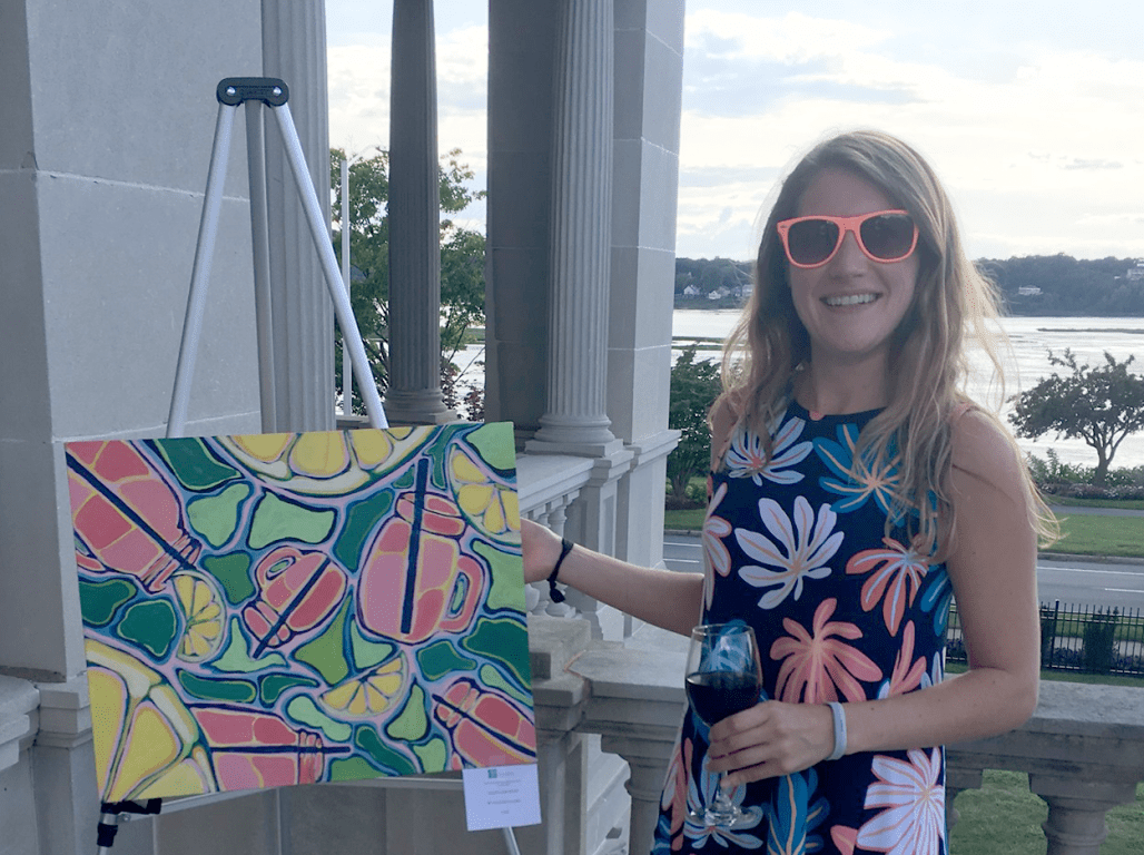 Allison Juliana wears neon sunglasses as she paints at an easel on a waterfront patio. 