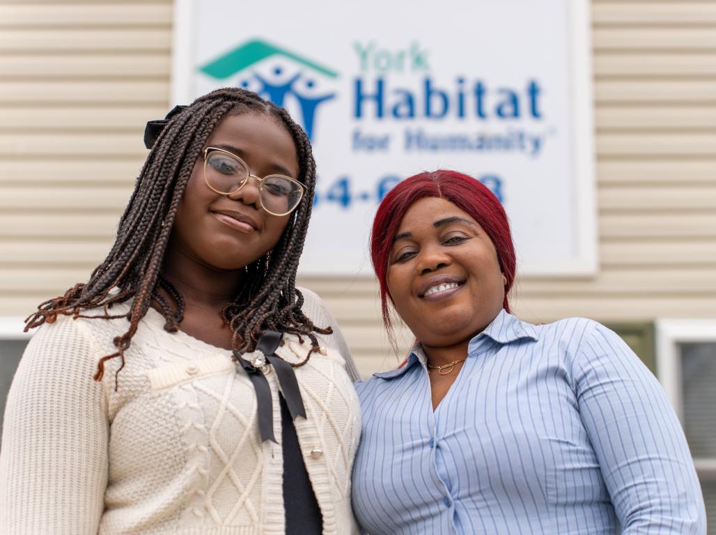 Elisee Okenge and her mother pose outside a house with a Habitat for Humanity banner hanging out front.