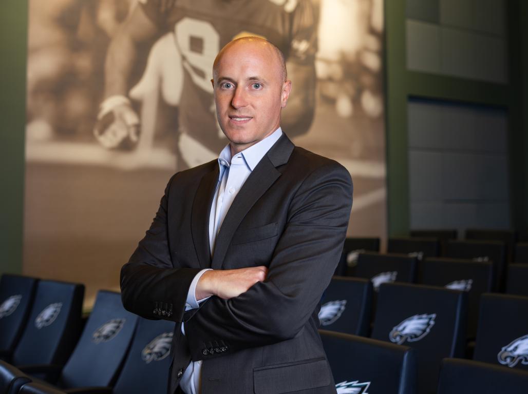 A person in business attire poses for a formal portrait with Philadelphia Eagles imagery visible.