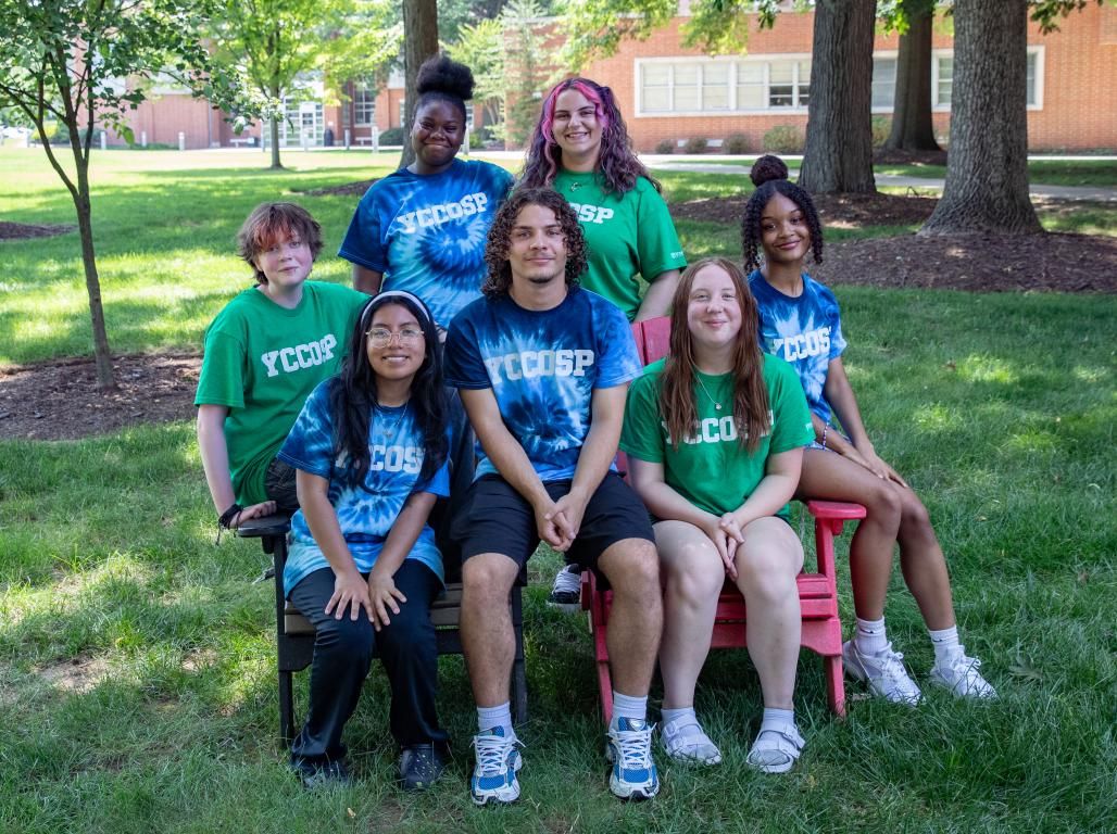 A group of college students pose for a photo outdoors on a sunny day. Many are wearing shirts with the letters YCCOSP.