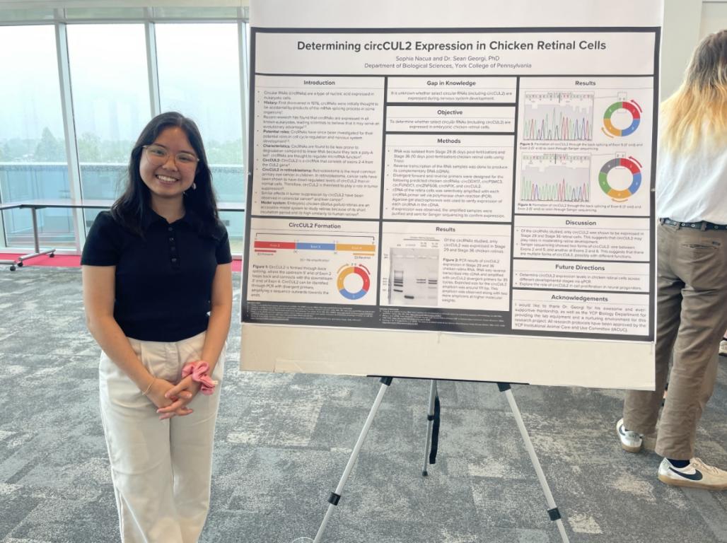 A college student poses in front of a research study poster at an event.