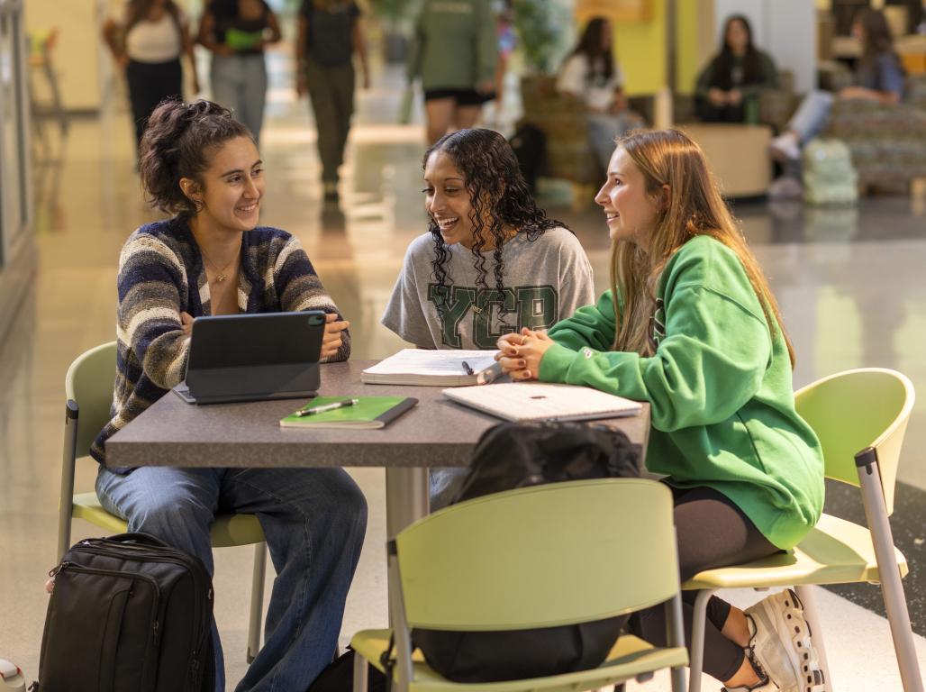 Three York College students at a round table in a busy building smile and talk. Notebooks, pens, and a tablet are visible.
