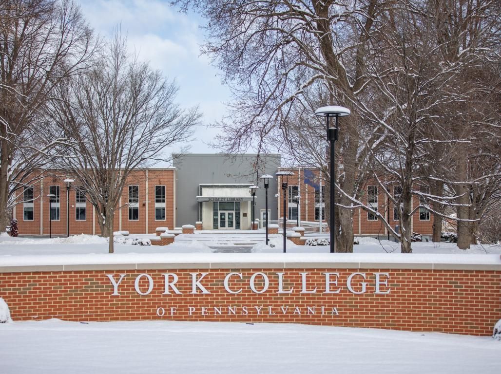 York College of Pennsylvania signage on brick entry to campus with the library building visible in the background. The ground is covered in snow.