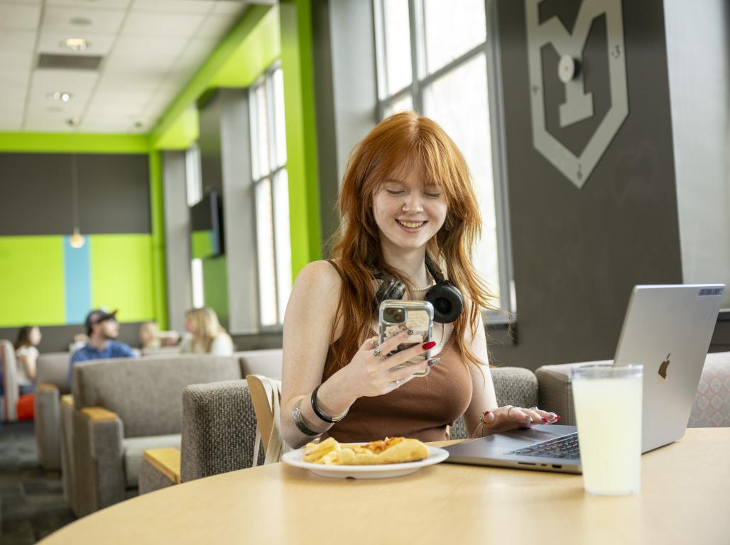 A college student smiles while looking at their phone in a dining hall. A laptop, plate of food, and glass of milk are visible on the table.