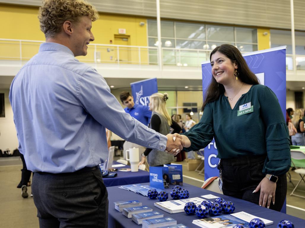 Smiling people shaking hands at a professional networking event, one is wearing a name tag with a green "alumni" label. A display table displaying promotional items and swag sits between them.