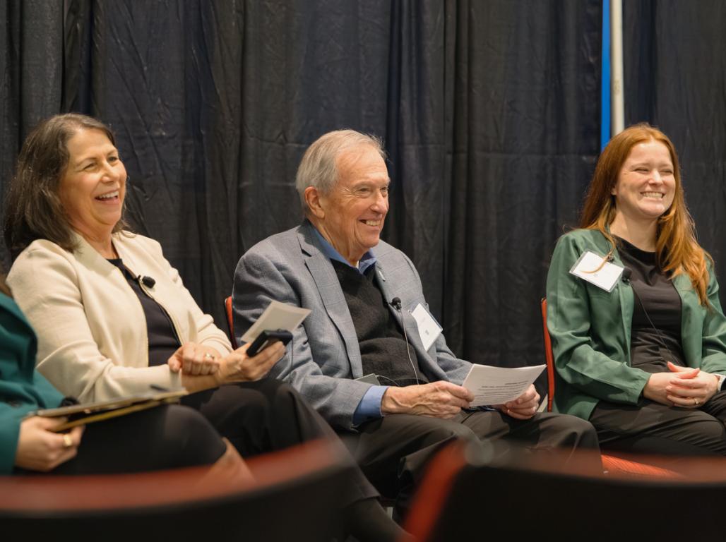 Three women and a man sit together smiling at an audience off camera. 