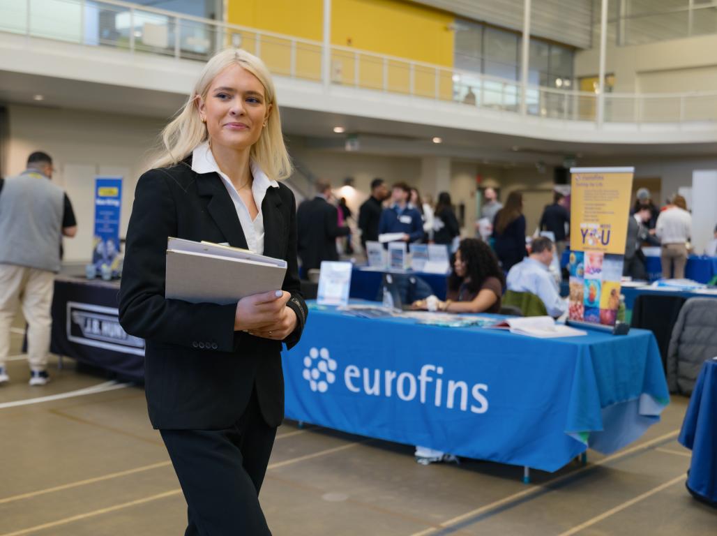 College student in business attire walks through a professional networking event, holding a folder.
