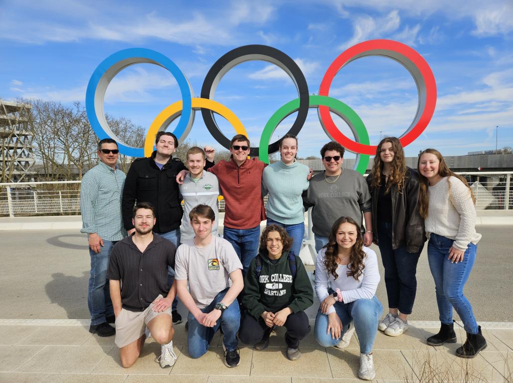 A group of York College students and faculty members pose together in front of a large Olympic rings display under a bright blue sky.