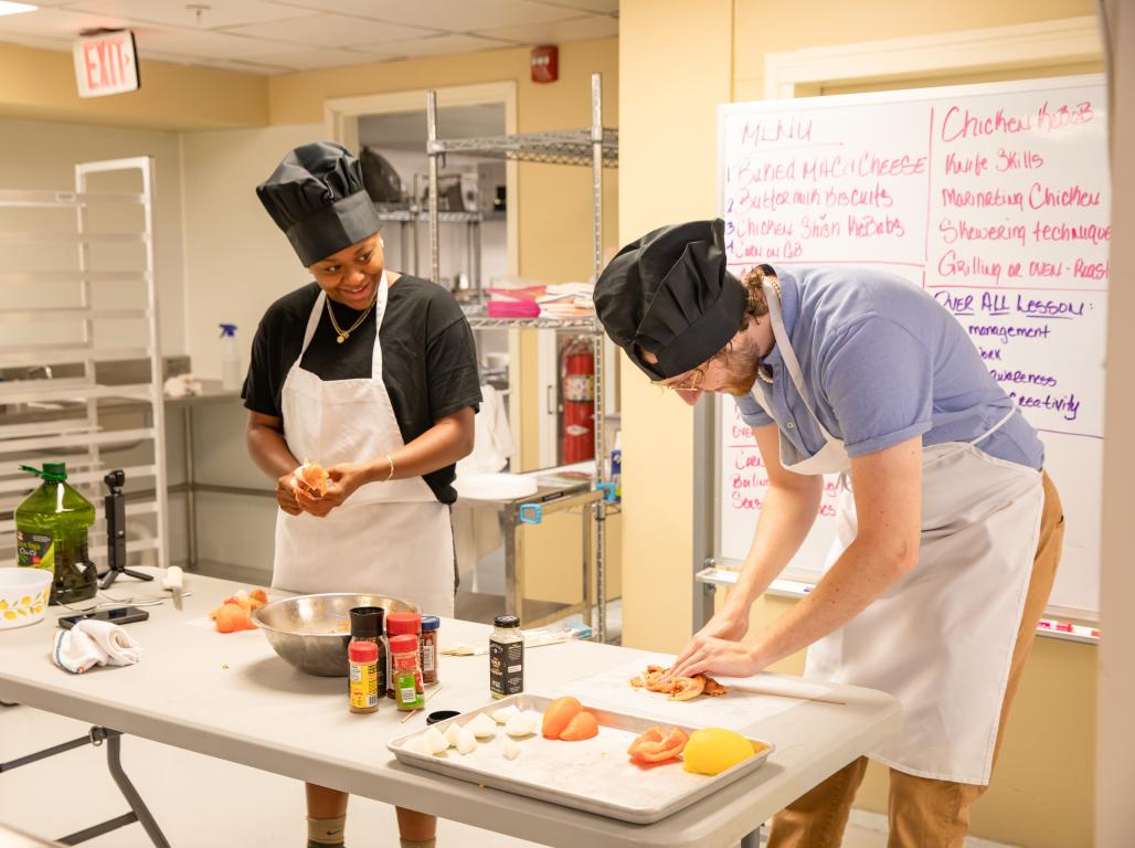 Two people prepare food in a kitchen wearing aprons and hats. A large whiteboard with writing is behind them.
