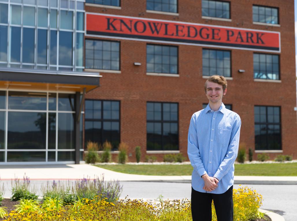 Christopher Magaha ’27 stands outside York College’s Knowledge Park building, with the brick-and-glass exterior, Knowledge Park sign, and landscaped flower beds behind him.