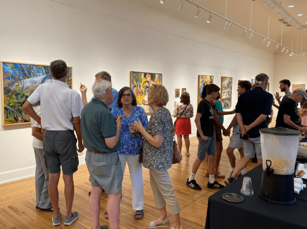 Community members gather and talk during an art exhibition reception at Marketview Arts, with paintings displayed along the gallery walls.